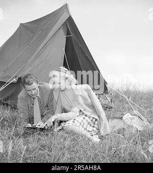 Camping in the old days. The young couple Barbro Santesson and Kurt Ferström sit outside their tent and carefully study a map and a compass. They have equipped themselves for outdoor life with a backpack and a fishing net lying in the grass. But they don't appear to be fully devoted outdoorsmen as he wears a tie and she a fashionable coat. Sweden 1945. Kristoffersson ref P46-1 Stock Photo