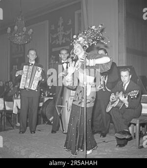 Young men in an orchestra and a singing young woman at the microphone perform on stage in a dance restaurant. The singer is exotically dressed with eye-catching clothing and headdress sprinkled with exotic fruits and flowers. Sweden 1945. Kristoffersson ref R61-6 Stock Photo