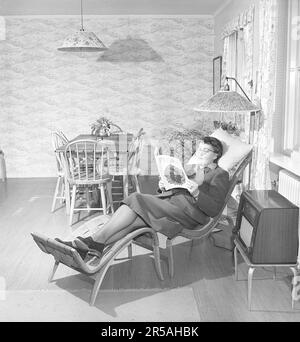 Home decor in the past. A living room where a woman lies in Bruno Mathsson's classic Pernilla lounge chair with footstool and reads a newspaper. On a table next to it is a radio. The lamps have pleated fabric shades and in the same room also a dining table with matching chairs. Sweden 1950. Kristoffersson ref AX79-3 Stock Photo