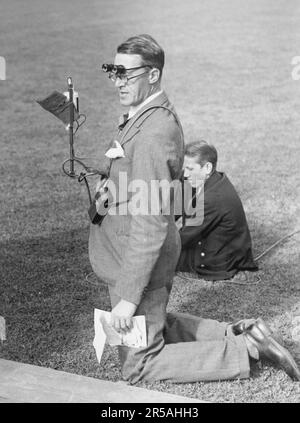 1940s media man.  A man is standing on his knees on the grass of the inner field of a sports stadium. He is radio reporter Sven Jerring 1895-1979 who reports from a sports event live on radio. The microphone is seen mounted in a stand hanging on his front. He was a very tall man so standing on his knees would still make him tall enough to see over the crowd... Sweden 1945 Stock Photo