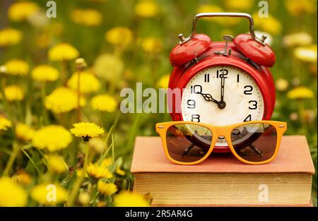 Alarm clock and sunglusses on a book with yellow flowers in the grass. End of school, summer break time. Stock Photo