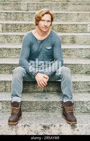 Outdoor fashion portrait of handsome red-haired man sitting on steps, wearing stylish blue pullover, grey jeans and brown boots Stock Photo