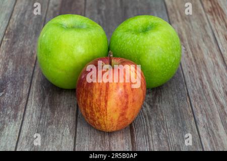 A three-piece still life composition featuring two green and one red apple sitting on a sliced wooden table Stock Photo