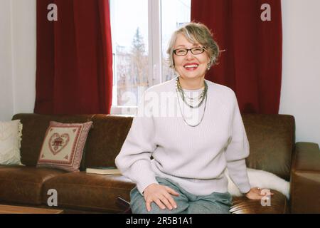 Interior portrait of 50 - 55 year old woman sitting on couch , wearing glasses Stock Photo