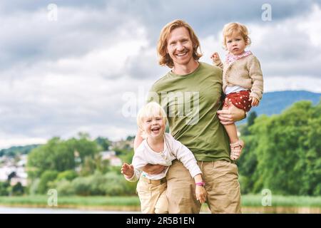 Outdoor portrait of happy young father holding two children in arms, toddler girl and preschooler boy having fun with dad on a nice sunny day Stock Photo