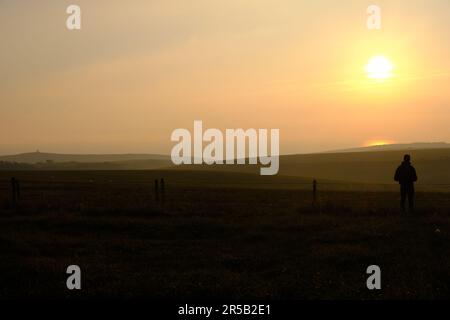 Eastbourne, East Sussex, United Kingdom - 9 October 2021. Man alone standing on hillside looking out across farmland towards a setting sun Copy space Stock Photo