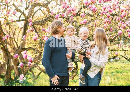 Outdoor portrait of happy young family playing in spring park under blooming magnolia tree, lovely couple with two little children having fun in sunny Stock Photo