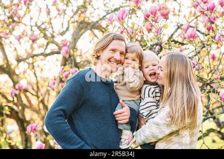 Outdoor portrait of happy young family playing in spring park under blooming magnolia tree, lovely couple with two little children having fun in sunny Stock Photo