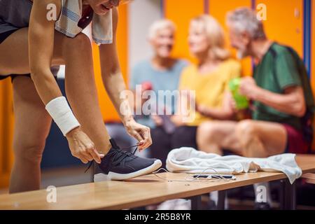 People in gym locker room. Young woman tying her shoe, seniors in the backgorund sitting and talking. Health, active lifestyle, wellness concept. Stock Photo