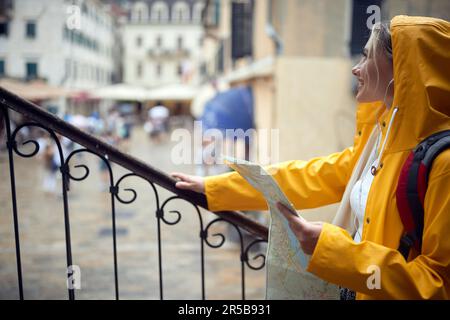 Travel in city in rain. Young woman in yellow raincoat hiding from rain, looking out and smiling. Stock Photo