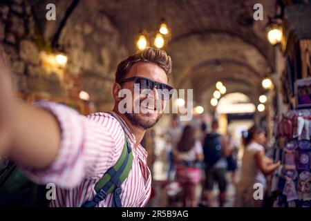 Young man taking selfie in a street alley shop bazar. Tourist on vacation walking through street bazar. Holiday, fun, shopping, lifestyle concept. Stock Photo