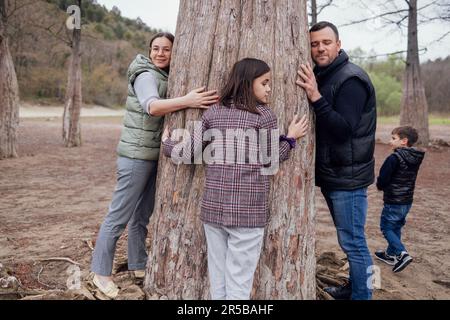 Young attractive woman, man and pretty teenage girl are hugging large cypress trunk. Happy family walks among trees. Little cute boy looks around. Pro Stock Photo