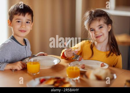 Two small children are sitting at dining table and having breakfast. Cute kids smile and look at camera. Lovely brother and sister in casual clothes a Stock Photo