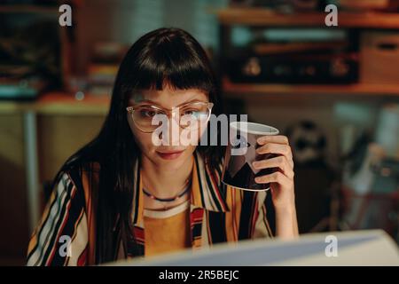 Young woman in eyeglasses drinking coffee and working over new project on computer Stock Photo