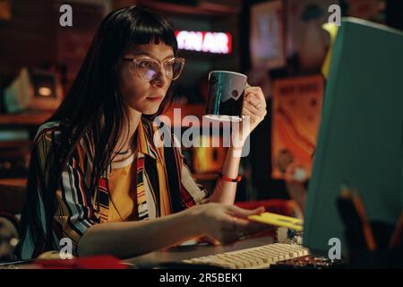Young woman in eyeglasses with cup of coffee using floppy disk to save information on computer while sitting at her workplace Stock Photo