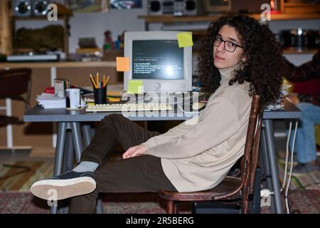 Portrait of young programmer with curly hair looking at camera sitting at his workplace with computer Stock Photo