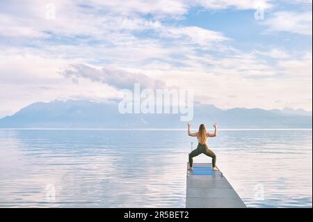 Outdoor portrait of young beautiful woman practicing yoga on dock by the lake, cactus pose, back view Stock Photo