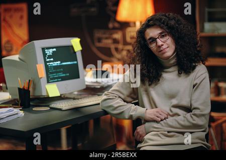 Portrait of young developer with long curly hair looking at camera while sitting at his workplace with computer and working with codes Stock Photo