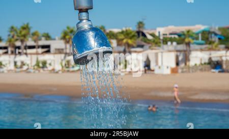 Outdoor refreshing shower head  spraying water to take shower on the pier. Blurry beach and swimming people in the background. Stock Photo