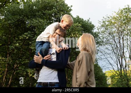 Father Carried Little Child, Son On Shoulder. Mother Walks Beside In Park. Summer Time. Happy Parenthood, Family Leisure Time. Love And Care Stock Photo