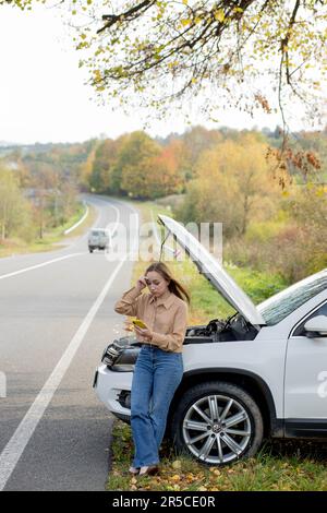 A woman waits for assistance near her car broken down on the road side. Woman with a broken car with open hood. Upset young woman with cell phone near broken car. Stock Photo