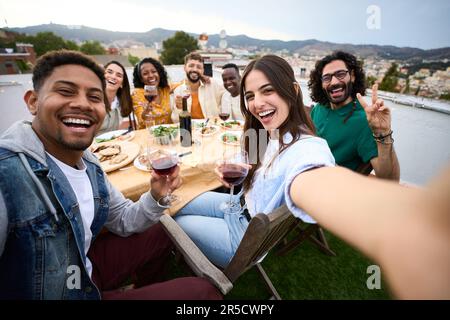 Group diverse friends having fun at rooftop party. Beautiful woman taking selfie at barbecue time.  Stock Photo