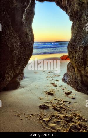 Footprints on sand leading to Pacific ocean through sea cave on Caves beach in Australia during sunrise. Stock Photo
