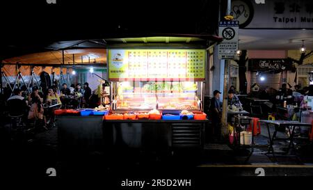 Street food stand at Huaxi Street Tourist Night Market in Taipei, Taiwan; tables and dining customers seated in background. Stock Photo