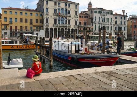 A tourist from behind sitting on the waterfront of the Grand Canal next to a cargo boat with working men, Venice, Veneto, Italy Stock Photo