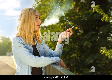 Woman smoking cigarette near railing outdoors on sunny day Stock Photo