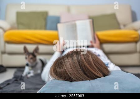 White woman lying on the floor while reading a book and resting next to her dog. Stock Photo