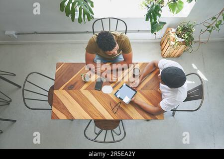 Startups require lots of coffee. High angle shot of two unrecognizable friends sitting together and using a tablet in a coffeeshop. Stock Photo