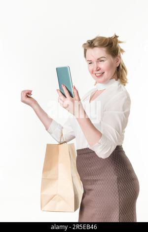 Middle aged adult woman holding shopping bags with purchases, smiling looking at phone, using app on smartphone. Caucasian ethnicity woman shopper Stock Photo
