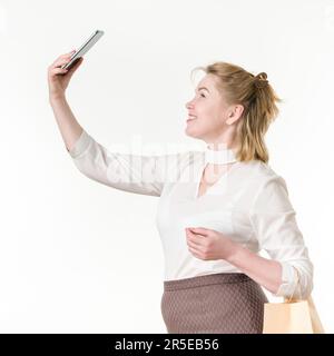 Caucasian ethnicity woman taking selfie holding phone up high, holding shopping bags with purchases. 49 years old smiling woman shopper Stock Photo