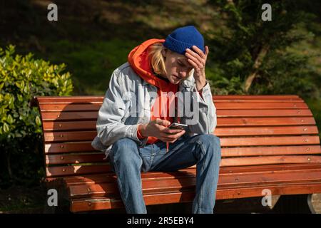 Upset millennial guy sitting on bench in park looking at smartphone, breaking up with girlfriend Stock Photo