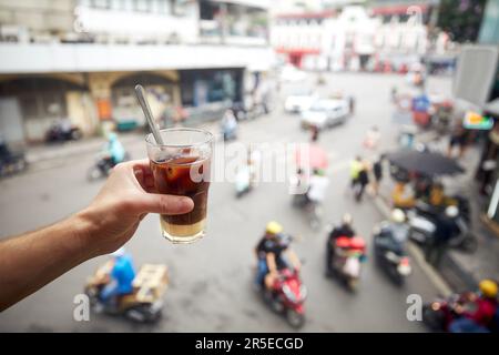 Hand holding of glass of Vietnamese iced coffee with condensed milk. Man drinking coffee above busy street of Old Quarter in Hanoi in Vietnam. Stock Photo