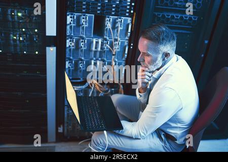 It takes a technical mind to solve this. a mature man looking thoughtful while using a laptop in a server room. Stock Photo