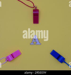 Three whistles of different colors arranged around the letter A on a yellow background. Minimalistic flat lay scene. Stock Photo