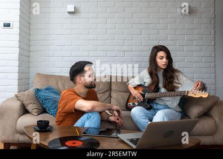 Teaching her to play guitar. Handsome young bearded man teaching his girlfriend to play guitar, sitting at home together. Musician couple enjoying fre Stock Photo