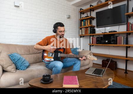 Young male musician beginner wearing modern headset learning to play guitar and record music at web video course using laptop. Concentrated millennial Stock Photo