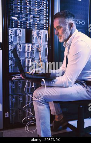 These machines have to stay running optimally. a mature man using a laptop while working in a server room. Stock Photo