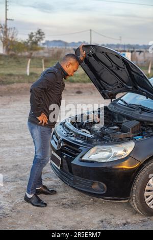 Young Latino man looking at the engine of his broken down car on the side of the road. Stock Photo