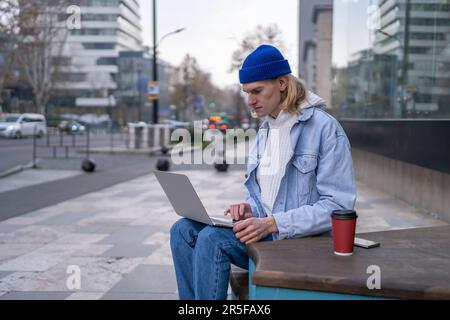 Millennial guy it developer sitting on empty city street with laptop working online Stock Photo