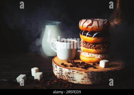 Stack of assorted donuts, a jug of milk and a cup of steaming coffee on a dark background. Stock Photo