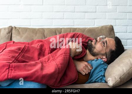 Absolute solitude. Side view of a depressed unhappy sad man lying on the sofa and hugging a blanket while feeling lonely, sad and miserable. Depressio Stock Photo
