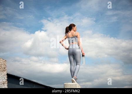 Rear view of a sporty woman runner holding a glass bottle of water. Sky background. Stock Photo