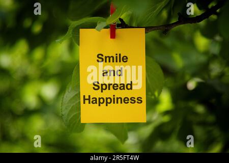 A yellow paper note with the words Smile and Spread Happiness on it attached to a tree branch with a clothes pin. Close up. Stock Photo