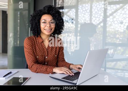 Portrait of mature adult successful businesswoman, african american woman at workplace smiling and looking at camera, confident boss working at workstation inside office, using laptop. Stock Photo