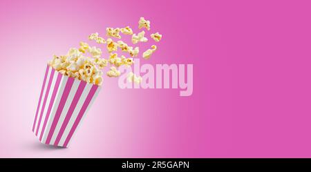 Popcorn flying out of pink white striped paper box isolated on pink background with copy space. Splash, levitation of popcorn grains. Stock Photo