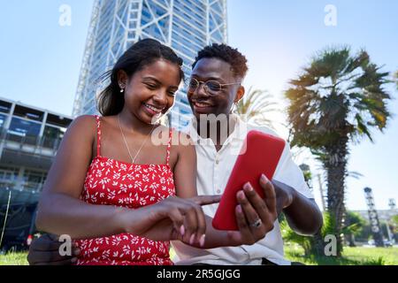 Young African American couple sitting outdoors using smart phone together on summer holidays. Stock Photo
